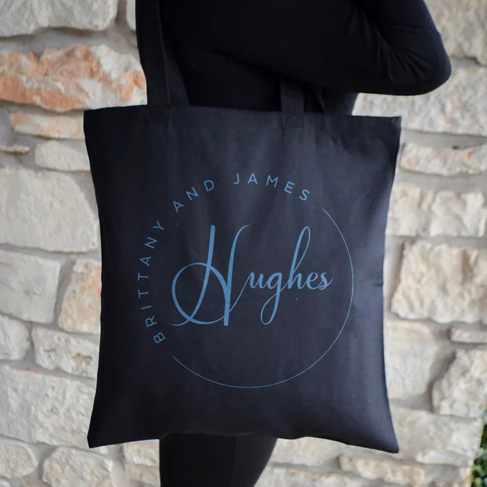 A person is holding a black tote bag with the text "Brittany and James Hughes" printed on it.