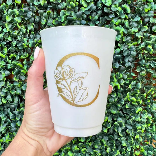 A hand holding a white cup with a gold letter "C" on it, against a background of green leaves.